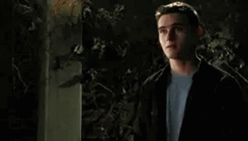 A Young Man In A Black Jacket And White Shirt Is Standing In Front Of A Tree At Night . GIF