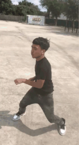 A Young Man In A Black Shirt And Grey Jeans Is Standing In A Parking Lot GIF