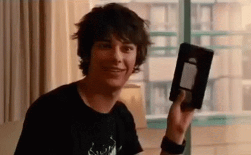 A Young Man In A Black Shirt Is Holding A Vhs Tape In His Hand . GIF