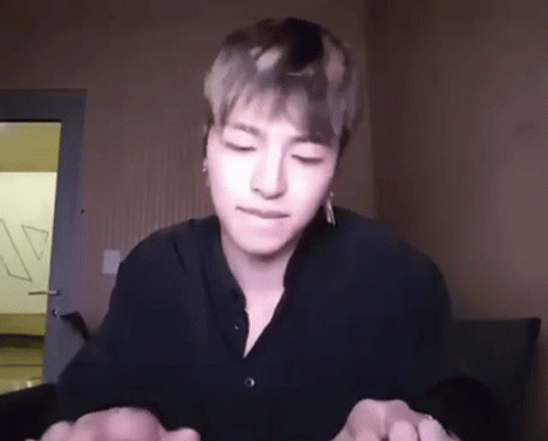 A Young Man In A Black Shirt Is Sitting In A Room With His Eyes Closed And Holding His Hands Together . GIF