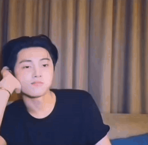 A Young Man In A Black Shirt Is Sitting On A Bed With His Hand On His Ear . GIF