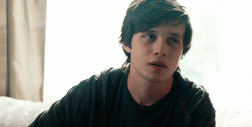 A Young Man In A Black Shirt Is Sitting On A Couch Looking At The Camera . GIF