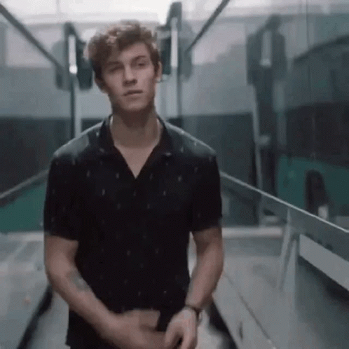 A Young Man In A Black Shirt Is Standing In A Hallway Looking At The Camera . GIF