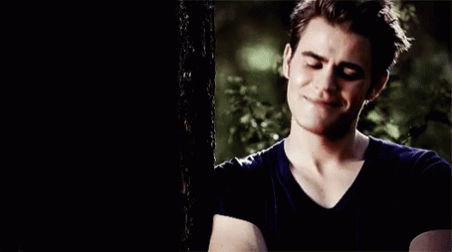 A Young Man In A Black Shirt Is Standing Next To A Tree And Smiling . GIF