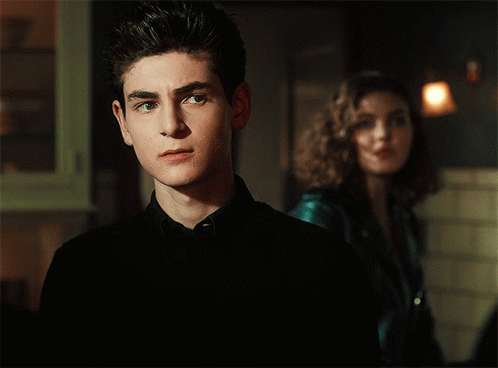 A Young Man In A Black Shirt Stands In Front Of A Woman In A Blue Jacket GIF