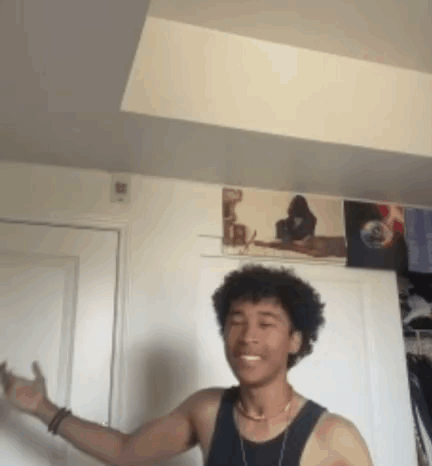 A Young Man In A Black Tank Top Is Standing In A Room With A Picture On The Wall Behind Him . GIF