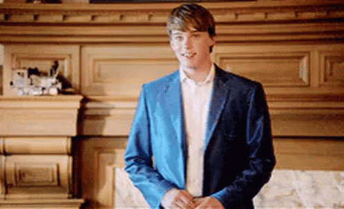 A Young Man In A Blue Jacket And White Shirt Is Standing In Front Of A Fireplace . GIF