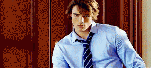 A Young Man In A Blue Shirt And Tie Is Standing In Front Of A Wooden Door . GIF