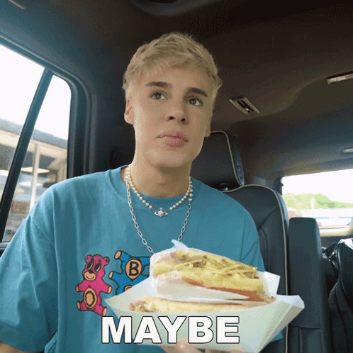 A Young Man In A Blue Shirt Is Holding A Sandwich And The Word Maybe Is Behind Him GIF