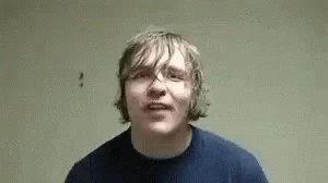 A Young Man In A Blue Shirt Is Making A Funny Face While Standing In Front Of A Wall . GIF