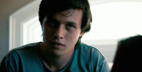 A Young Man In A Blue Shirt Is Sitting In Front Of A Window Looking At Something . GIF