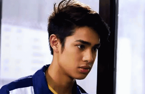 A Young Man In A Blue Shirt Is Standing Next To A Window And Looking At The Camera . GIF