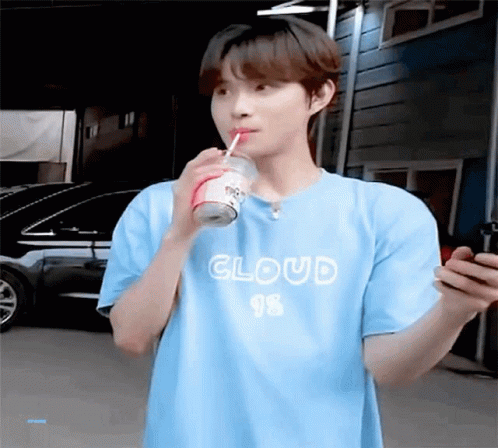 A Young Man In A Blue T-Shirt Is Drinking A Soda Through A Straw While Looking At His Phone . GIF