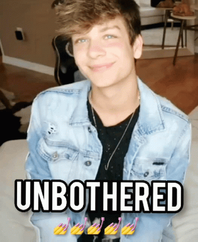 A Young Man In A Denim Jacket Is Sitting On A Couch With The Words " Unbothered " Below Him GIF