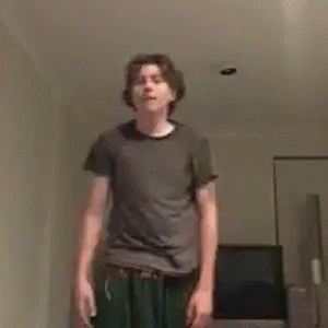 A Young Man In A Gray Shirt And Green Pants Is Standing In A Living Room . GIF