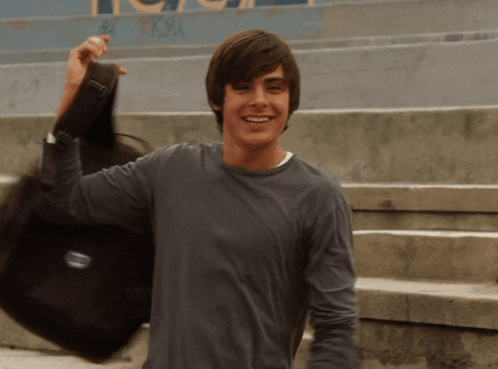 A Young Man In A Gray Shirt Is Holding A Black Bag With The Letter T On It GIF