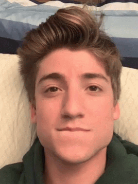 a young man in a green hoodie is laying on a bed and making a funny face
