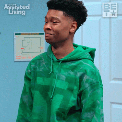 A Young Man In A Green Hoodie Is Standing In Front Of A Sign That Says " Assisted Living " GIF