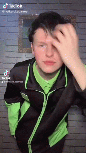 A Young Man In A Green Shirt And Black Jacket Is Standing In Front Of A Brick Wall . GIF