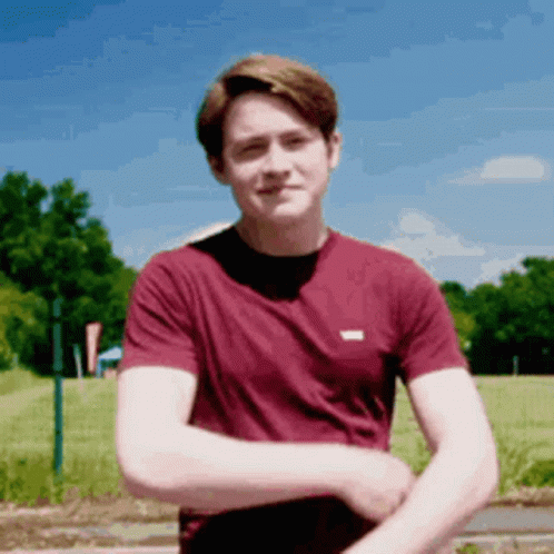 A Young Man In A Maroon Shirt Is Standing In A Field With His Arms Crossed . GIF