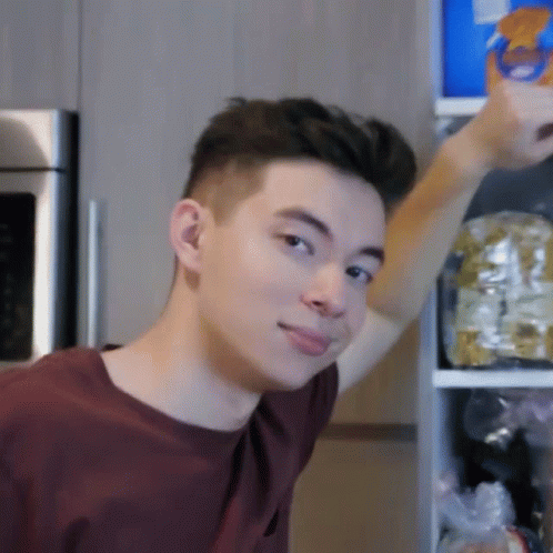 A Young Man In A Maroon Shirt Is Standing In Front Of A Refrigerator Reaching For Something GIF