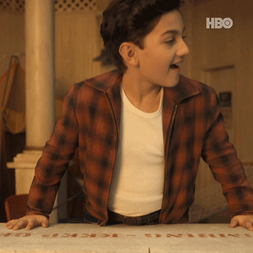 A Young Man In A Plaid Jacket Is Standing Next To A Box That Says Hbo On It GIF