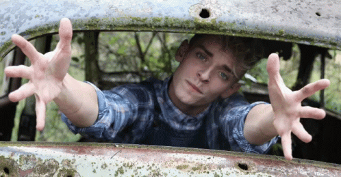 a young man in a plaid shirt is reaching out from a rusted car window