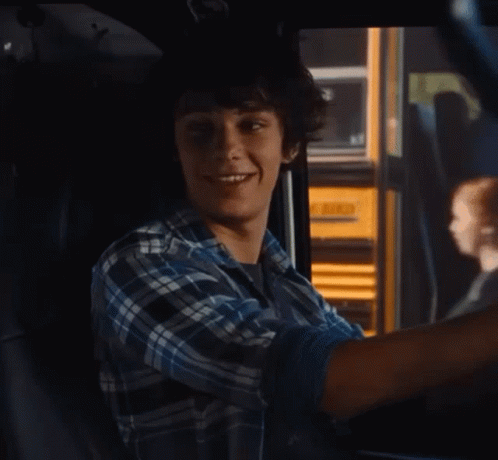 A Young Man In A Plaid Shirt Is Sitting In A Car With A Yellow School Bus In The Background GIF