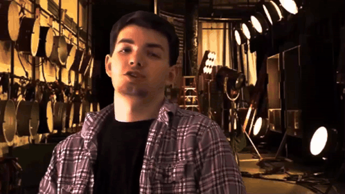 A Young Man In A Plaid Shirt Stands In Front Of A Wall Of Drums GIF