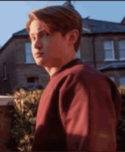 A Young Man In A Red Sweater Is Standing In Front Of A House And Looking At The Camera . GIF