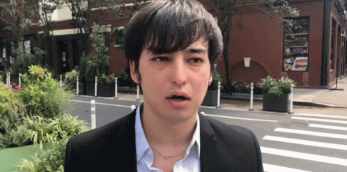 A Young Man In A Suit And Blue Shirt Is Standing In Front Of A Building . GIF