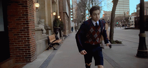 A Young Man In A Suit And Tie Is Running Down A Sidewalk In A City . GIF