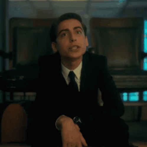 A Young Man In A Suit And Tie Is Sitting On A Chair . GIF