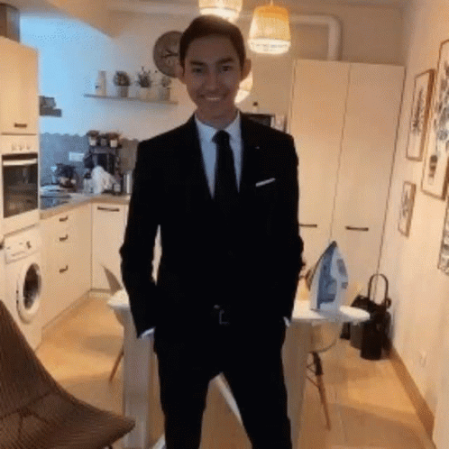 A Young Man In A Suit And Tie Is Standing In A Kitchen With His Hands In His Pockets . GIF