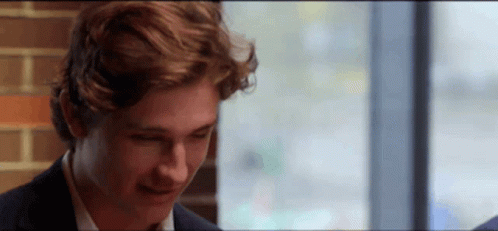 A Young Man In A Suit Is Sitting In Front Of A Window And Smiling . GIF