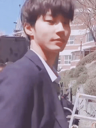 A Young Man In A Suit Is Standing In Front Of A Building Looking At The Camera . GIF
