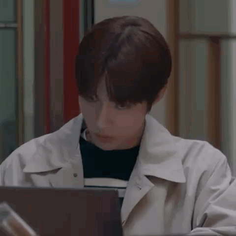 A Young Man In A Trench Coat Is Sitting In Front Of A Laptop . GIF