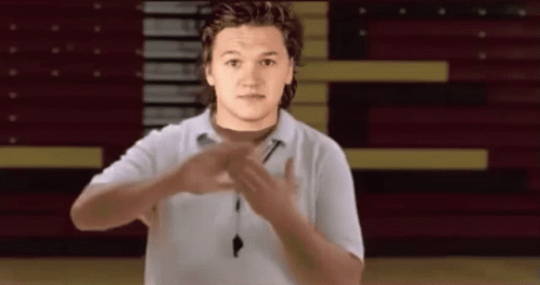 A Young Man In A White Shirt Is Making A Gesture With His Hands In Front Of A Basketball Court . GIF