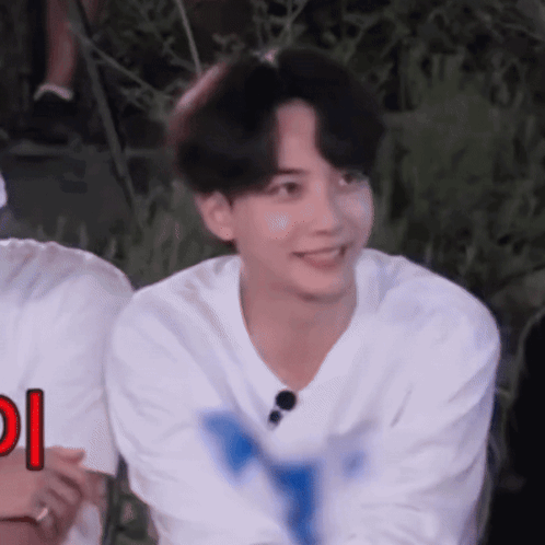 A Young Man In A White Shirt Is Smiling And Looking At The Camera While Sitting In A Chair . GIF
