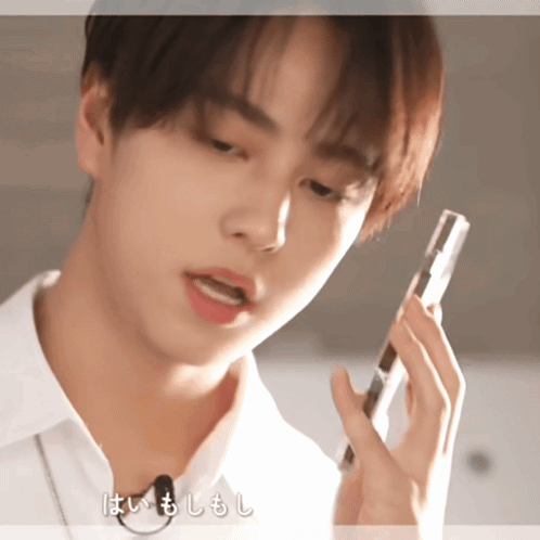 A Young Man In A White Shirt Is Talking On A Cell Phone With Chinese Writing Behind Him GIF