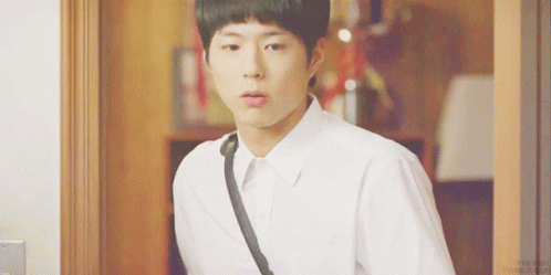 A Young Man In A White Shirt With A Black Strap Around His Neck Is Standing In Front Of A Door GIF