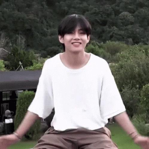 A Young Man In A White T-Shirt And Brown Shorts Is Sitting On A Lawn With His Arms Outstretched . GIF
