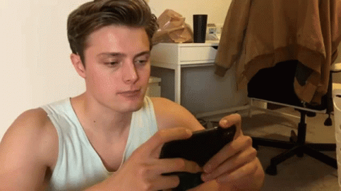 A Young Man In A White Tank Top Is Looking At His Phone GIF