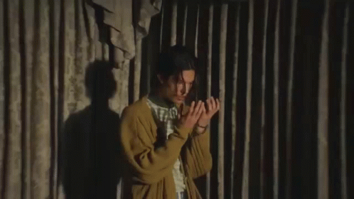 a young man in a yellow cardigan is making a funny face while sitting in front of a curtain .