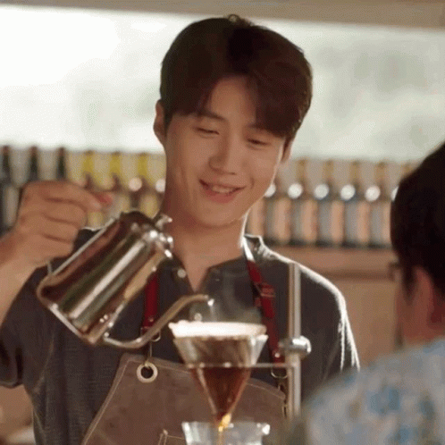 A Young Man In An Apron Is Pouring Coffee Into A Glass . GIF