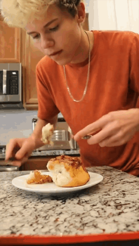 A Young Man In An Orange Shirt Is Eating A Cinnamon Roll GIF