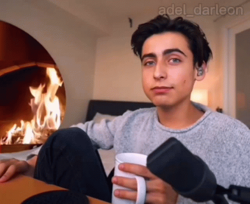 A Young Man Is Holding A Cup Of Coffee In Front Of A Fireplace GIF