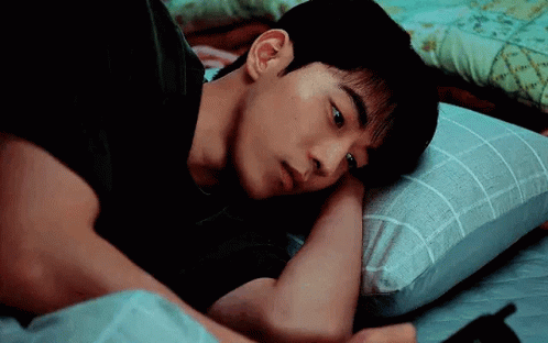 A Young Man Is Laying In Bed With A Pillow And Looking At His Phone . GIF