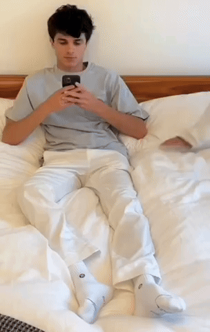 A Young Man Is Laying On A Bed Looking At His Cell Phone GIF