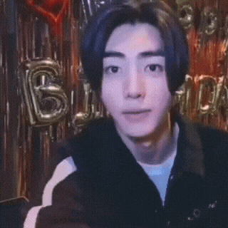 A Young Man Is Pointing At The Camera With Balloons Behind Him That Say Birthday GIF